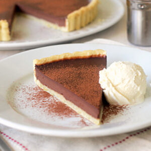 A slice of chocolate tart dusted with cocoa powder is served on a white plate with a scoop of vanilla ice cream. Another tart is visible in the background.