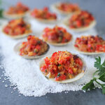 A close-up of stuffed clams topped with a colorful mixture of breadcrumbs, herbs, and diced vegetables, arranged on a bed of coarse salt with fresh parsley garnishes.