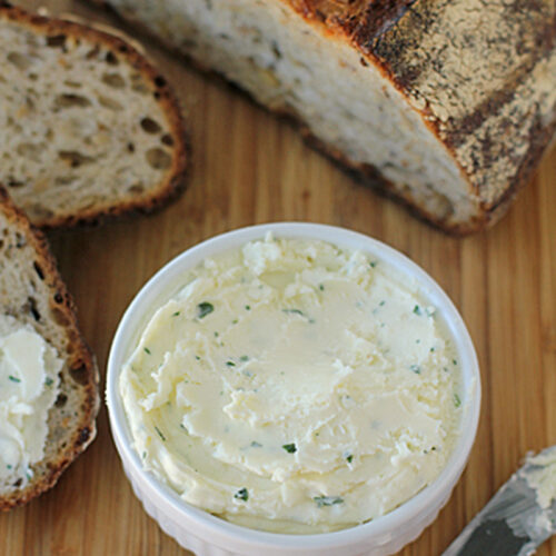 A white ramekin filled with herbed butter sits on a wooden surface, surrounded by slices of rustic bread, with a butter knife resting nearby.