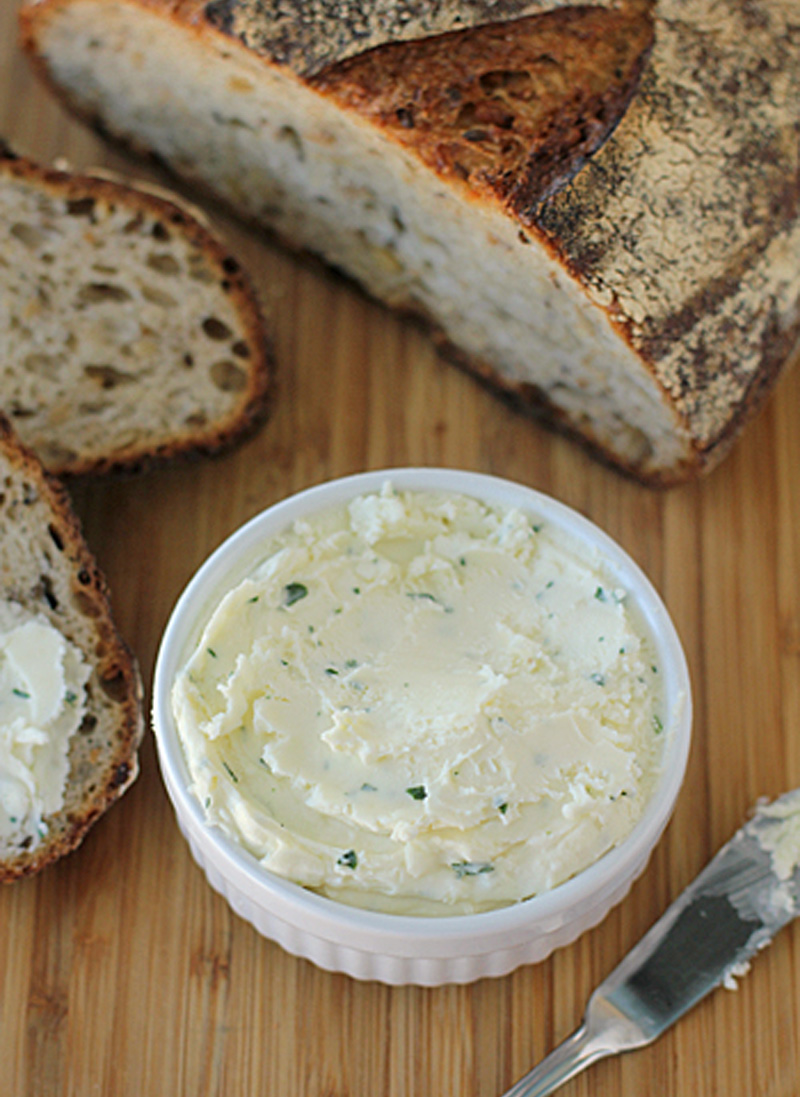 A white ramekin filled with herbed butter sits on a wooden surface, surrounded by slices of rustic bread, with a butter knife resting nearby.