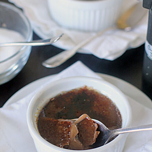 A white ramekin filled with cr&egrave;me br&ucirc;l&eacute;e sits on a napkin, with a spoon breaking through the caramelized sugar top. Another ramekin, a bowl of sugar, and a kitchen torch are visible in the background.