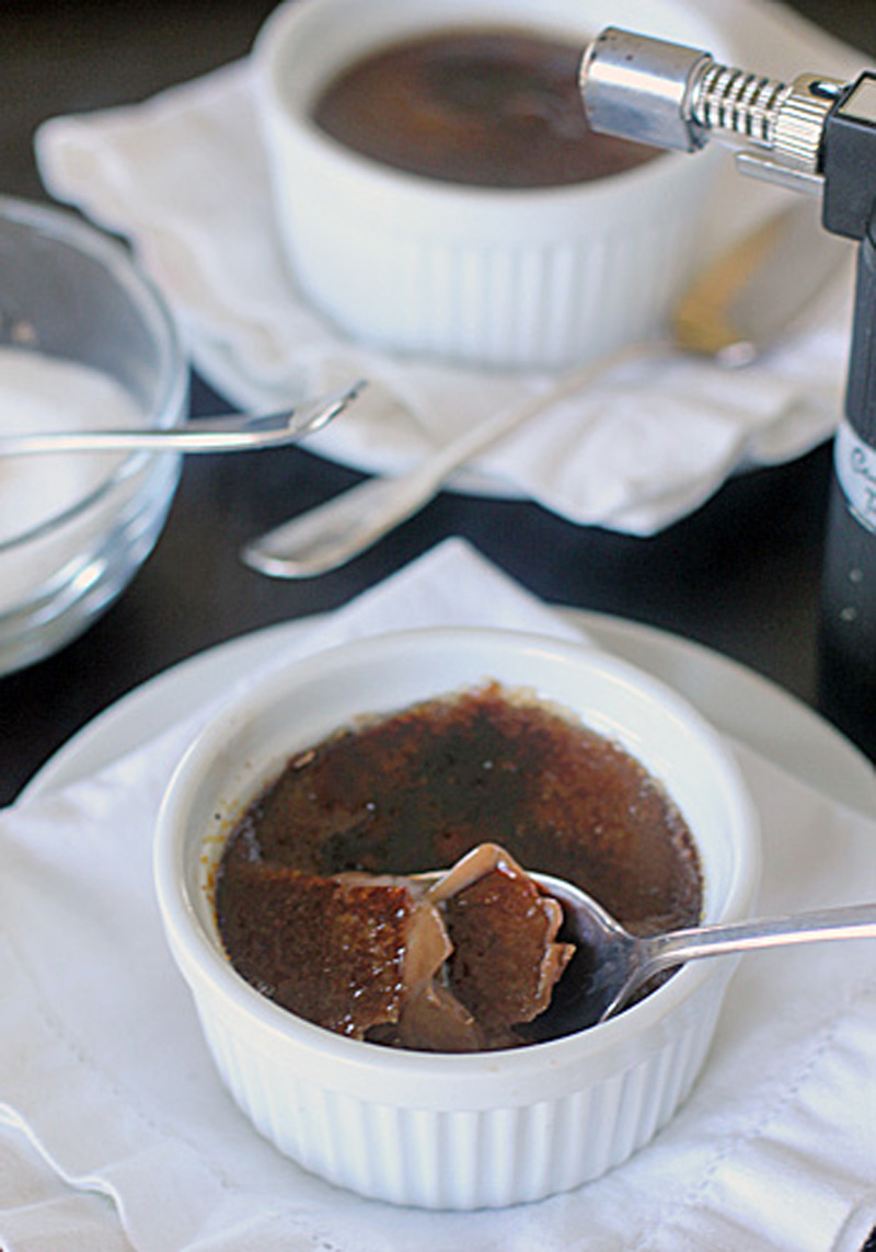 A white ramekin filled with crème brûlée sits on a napkin, with a spoon breaking through the caramelized sugar top. Another ramekin, a bowl of sugar, and a kitchen torch are visible in the background.