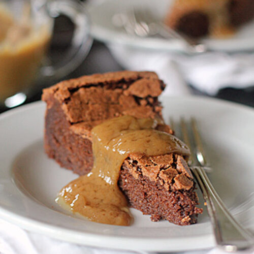 A slice of chocolate cake with a crumbly top is served on a white plate, topped with a generous drizzle of creamy sauce. A fork rests beside the cake, and another plate is blurred in the background.