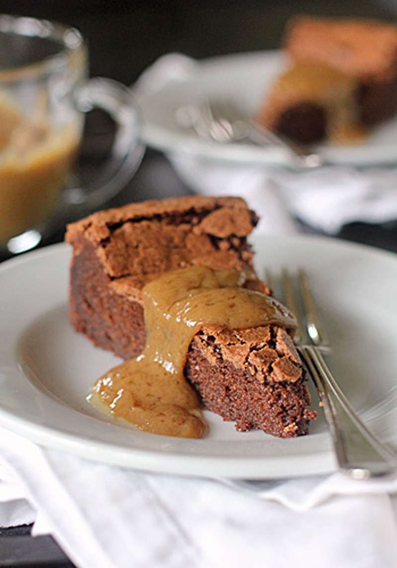 A slice of chocolate cake with a crumbly top is served on a white plate, topped with a generous drizzle of creamy sauce. A fork rests beside the cake, and another plate is blurred in the background.