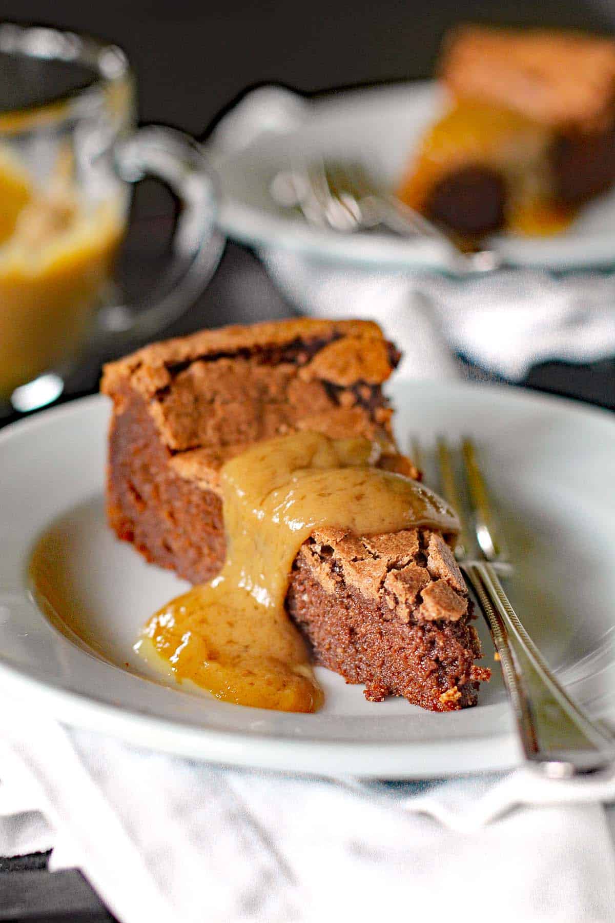 A slice of chocolate cake topped with a thick caramel sauce sits on a white plate with a fork, with a blurred cup and another plate in the background.