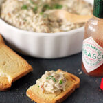 A small piece of toast topped with creamy dip sits on a dark surface next to a bottle of Tabasco sauce, with a bowl of dip and more toast in the background.