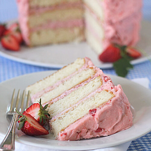 A slice of layered vanilla cake with pink strawberry frosting sits on a plate with a fork and fresh strawberry, with the rest of the cake in the background on a blue checkered tablecloth.