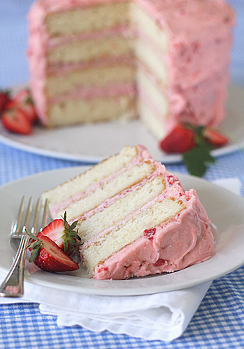 A slice of layered vanilla cake with pink strawberry frosting sits on a plate with a fork and fresh strawberry, with the rest of the cake in the background on a blue checkered tablecloth.