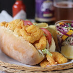 A shrimp po’ boy sandwich with lettuce and tomato on a hoagie roll sits on a plate with coleslaw, lemon wedges, and potato chips. Mardi Gras beads, hot sauce, beer, and soft drink bottles are in the background.