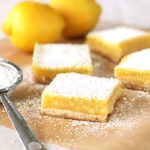 Four lemon bars dusted with powdered sugar are arranged on parchment paper, with a metal sifter and two whole lemons in the background. The lemon bars have a yellow filling and a light crust.