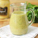 A clear glass pitcher filled with creamy, yellow-green salad dressing sits on a white cloth. In the background, there is a jar of mustard and a bunch of fresh parsley on a wooden surface.
