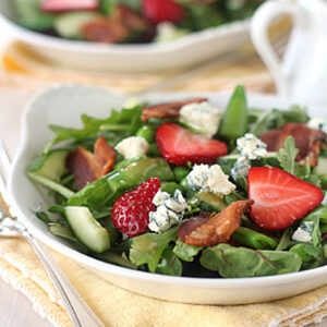 A fresh salad with arugula, sliced strawberries, crumbled blue cheese, cucumber, and pieces of bacon served in a white bowl on a yellow napkin, with another similar bowl in the background.