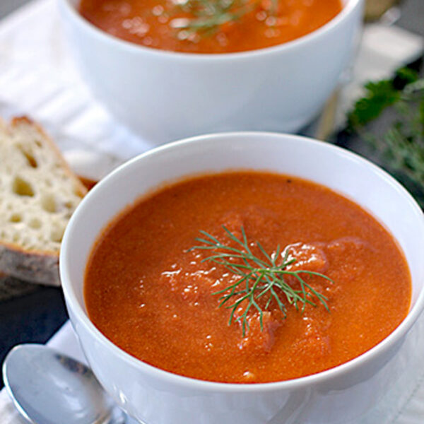 Two white bowls filled with tomato soup, each garnished with a sprig of fresh dill. A slice of rustic bread and a spoon are placed next to the bowls on a white napkin.