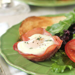 A breakfast plate with a baked egg in a ham cup, garnished with herbs, served alongside toast, mixed greens, and sliced tomato on a green plate.