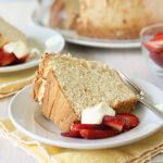 A slice of sponge cake served on a white plate with fresh sliced strawberries and a dollop of whipped cream, placed on a yellow napkin. Another plate and a bowl of strawberries are in the background.