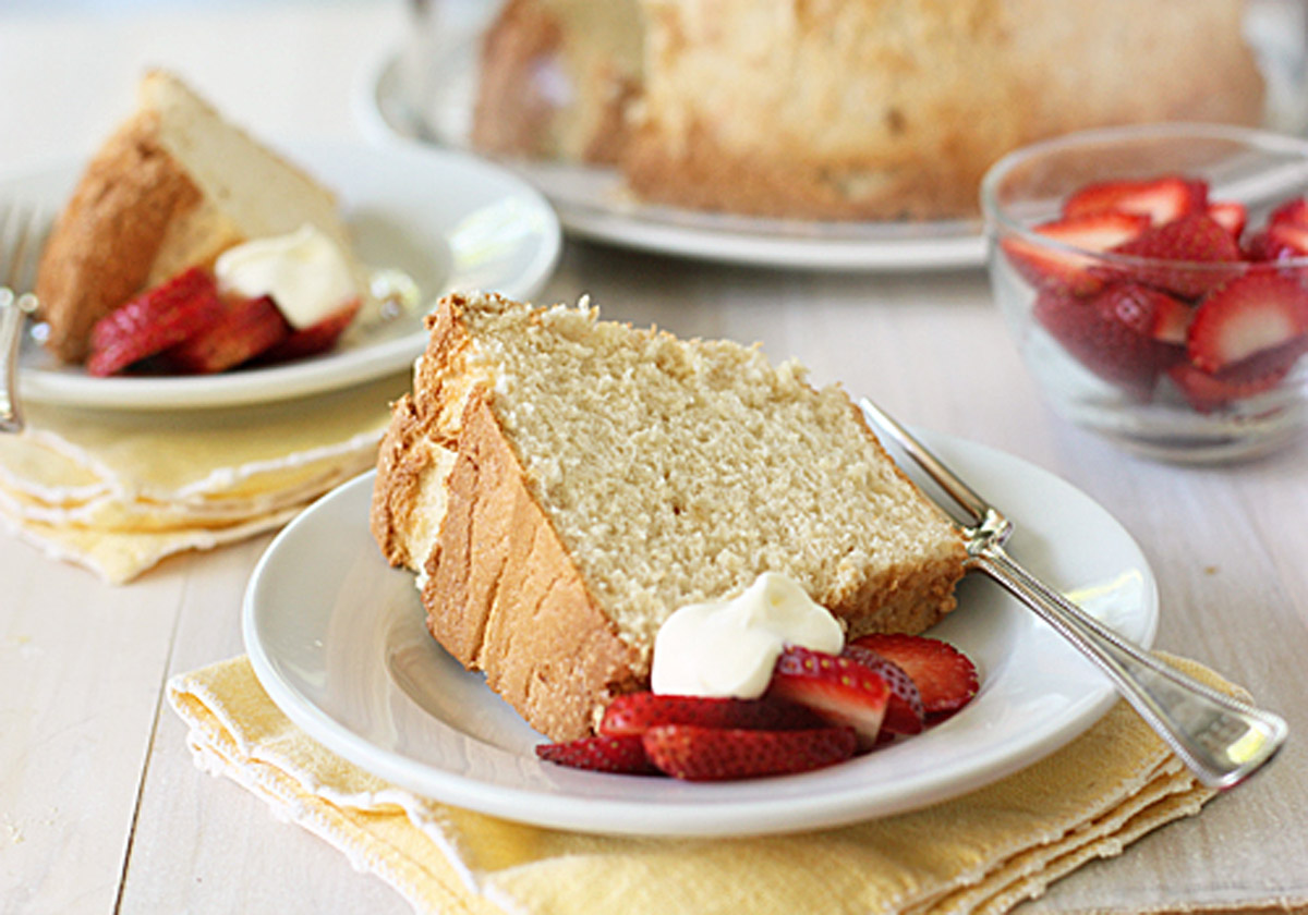 A slice of sponge cake served on a white plate with fresh sliced strawberries and a dollop of whipped cream, placed on a yellow napkin. Another plate and a bowl of strawberries are in the background.