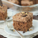 A slice of chocolate cake with chocolate frosting and chopped nuts on top, served on a white plate with a fork. Another slice and a glass baking dish with more cake are visible in the background.