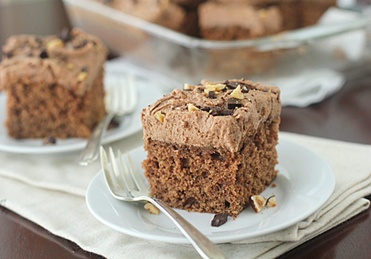 A slice of chocolate cake with chocolate frosting and chopped nuts on top, served on a white plate with a fork. Another slice and a glass baking dish with more cake are visible in the background.