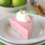 A slice of pink pie with a crumbly crust, topped with whipped cream and lime zest, sits on a white plate with a green napkin. In the background, whole limes and forks are slightly out of focus.