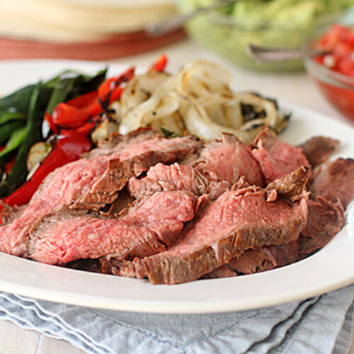 A plate of sliced grilled steak served with sautéed onions, red bell peppers, and green vegetables, sitting on a folded blue napkin, with guacamole and salsa in the background.