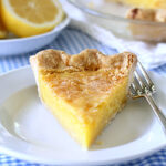 A slice of lemon pie with a golden, flaky crust sits on a white plate with a fork. In the background, the rest of the pie and a bowl with a halved lemon are visible on a blue and white checkered tablecloth.