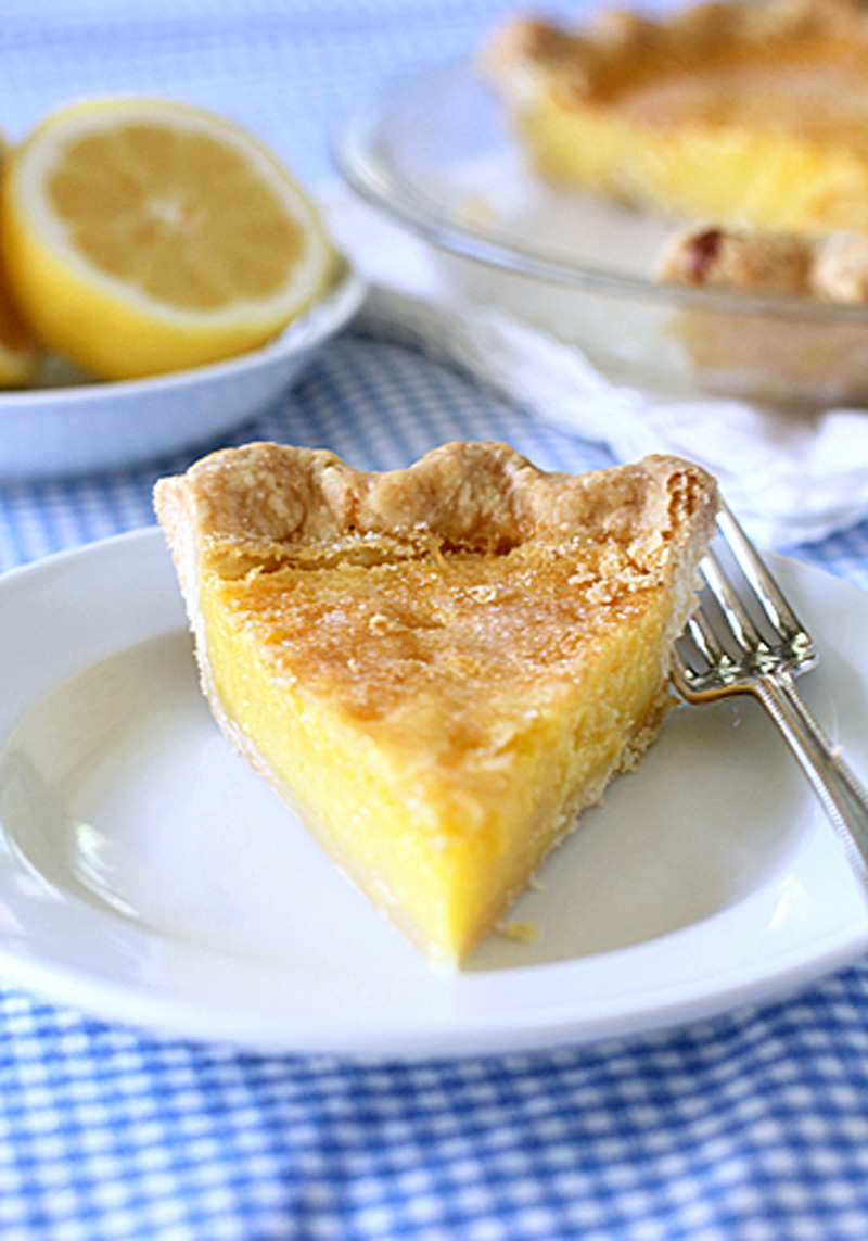 A slice of lemon pie with a golden, flaky crust sits on a white plate with a fork. In the background, the rest of the pie and a bowl with a halved lemon are visible on a blue and white checkered tablecloth.