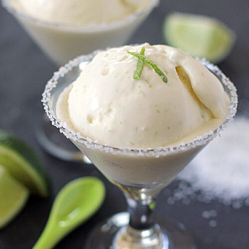 A martini glass filled with a scoop of creamy lime ice cream, garnished with lime zest and a salted rim. A green spoon and lime slices sit nearby on a dark surface. Another serving is blurred in the background.