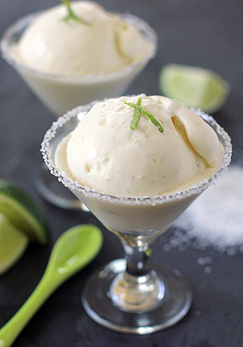 A martini glass filled with a scoop of creamy lime ice cream, garnished with lime zest and a salted rim. A green spoon and lime slices sit nearby on a dark surface. Another serving is blurred in the background.