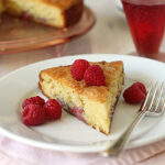 A slice of golden cake with raspberries on top and on the side, served on a white plate with a fork. A glass of raspberry drink and the rest of the cake are visible in the background.