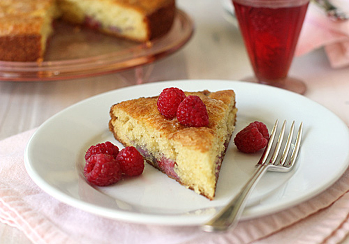 A slice of golden cake with raspberries on top and on the side, served on a white plate with a fork. A glass of raspberry drink and the rest of the cake are visible in the background.