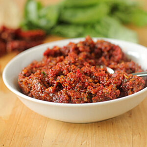 A white bowl filled with chunky red pesto sits on a wooden surface, with a spoon in the bowl and fresh basil leaves and sun-dried tomatoes in the background.