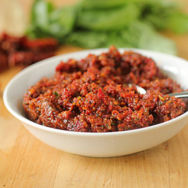 A white bowl filled with chunky red pesto sits on a wooden surface, with a spoon in the bowl and fresh basil leaves and sun-dried tomatoes in the background.