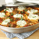 A skillet filled with lasagna noodles, tomato sauce, sausage, and large dollops of ricotta cheese, garnished with fresh basil, resting on a blue cloth on a wooden surface.