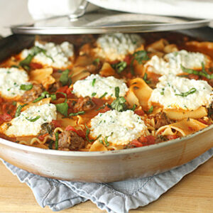 A skillet filled with lasagna noodles, tomato sauce, sausage, and large dollops of ricotta cheese, garnished with fresh basil, resting on a blue cloth on a wooden surface.