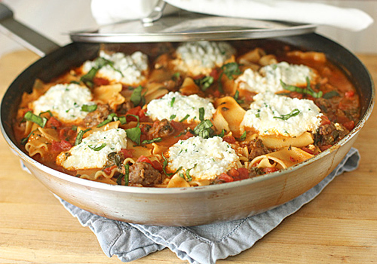 A skillet filled with lasagna noodles, tomato sauce, sausage, and large dollops of ricotta cheese, garnished with fresh basil, resting on a blue cloth on a wooden surface.
