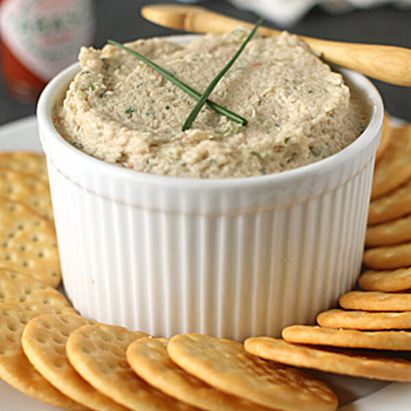 A white ramekin filled with a creamy dip, garnished with chives, is placed on a plate surrounded by round crackers. A small wooden spreader rests on the ramekin, and a bottle of Tabasco sauce is in the background.