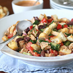 A large white bowl of salad with chunks of bread, tomatoes, cucumbers, olives, chicken, and herbs sits on a blue cloth. A spoon rests on the bowl, with plates, forks, and a bowl of dressing in the background.