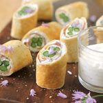 Sliced rolled crepes filled with cream cheese, asparagus, and herbs are arranged on a wooden board with a glass of dipping sauce and purple edible flowers scattered around.