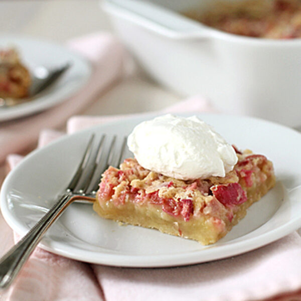 A slice of dessert bar topped with whipped cream sits on a white plate with a fork, resting on a pale pink napkin. Another plate and a baking dish can be seen in the background.