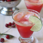 A glass of bright red drink with ice, garnished with a lime slice, sits on a white surface. Another similar glass and a metal bowl of cherries are blurred in the background. Loose cherries are scattered nearby.