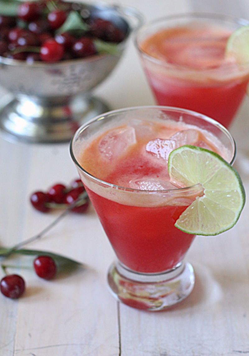 A glass of bright red drink with ice, garnished with a lime slice, sits on a white surface. Another similar glass and a metal bowl of cherries are blurred in the background. Loose cherries are scattered nearby.