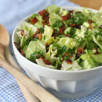 A white bowl filled with chopped lettuce, crispy bacon bits, and fresh herbs sits on a blue and white checkered cloth next to two wooden serving spoons.