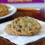 Two chocolate chip cookies rest on a white napkin, with a glass of milk, a pencil, and a textbook blurred in the background on a wooden table.