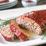 Sliced meatloaf with a shiny glaze served on a white platter, garnished with fresh thyme sprigs. A fork is placed beside the meatloaf and a small dish is visible in the background.