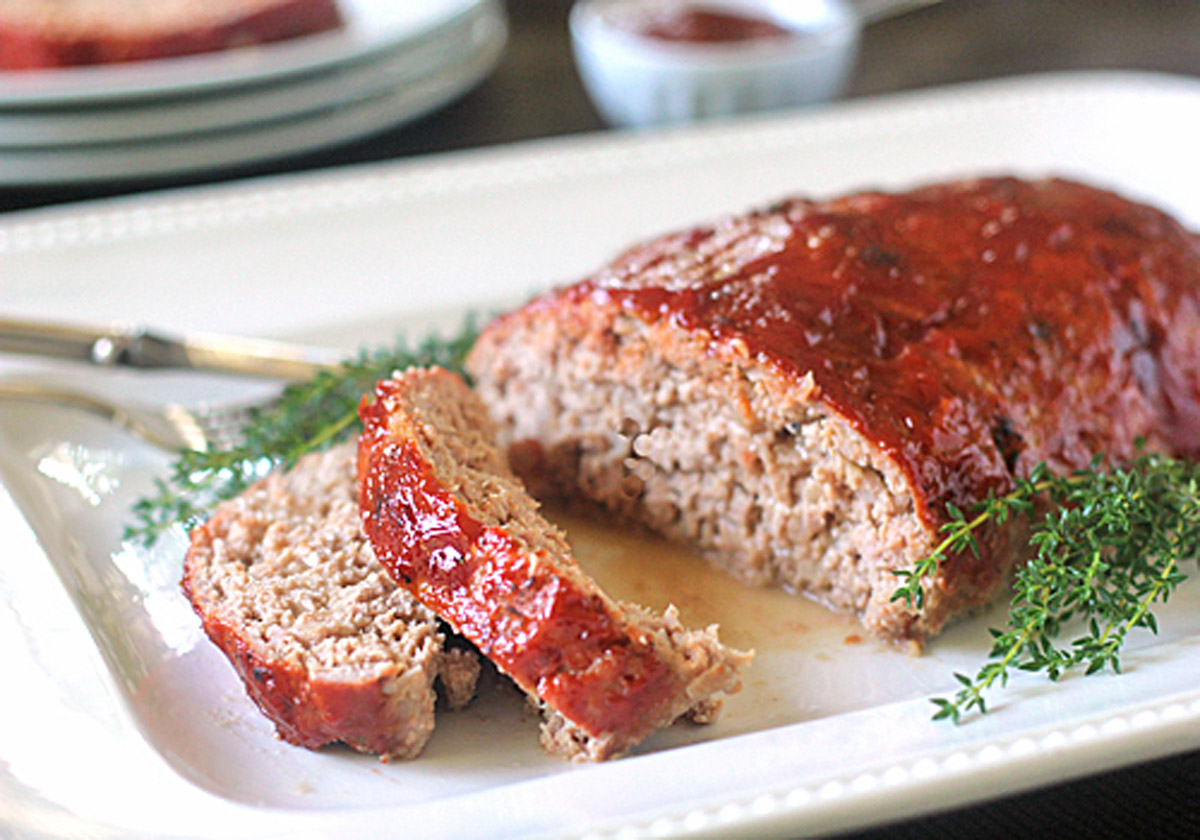 Sliced meatloaf with a shiny glaze served on a white platter, garnished with fresh thyme sprigs. A fork is placed beside the meatloaf and a small dish is visible in the background.