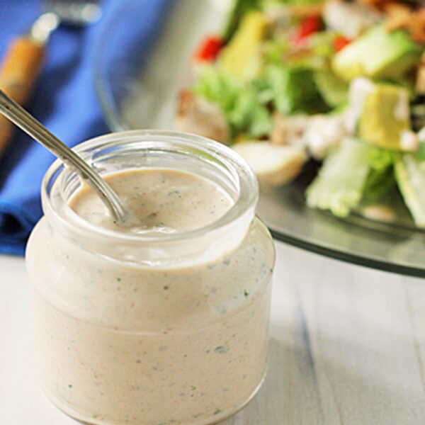 A small glass jar filled with creamy ranch dressing and a spoon, placed in front of a plate of fresh salad with leafy greens and vegetables. A blue napkin and fork are in the background.