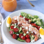 A salad with shrimp, cherry tomatoes, avocado, cucumber, bacon, feta cheese, and a halved boiled egg on a white plate. A small pitcher of pink dressing sits nearby on a blue checkered tablecloth.