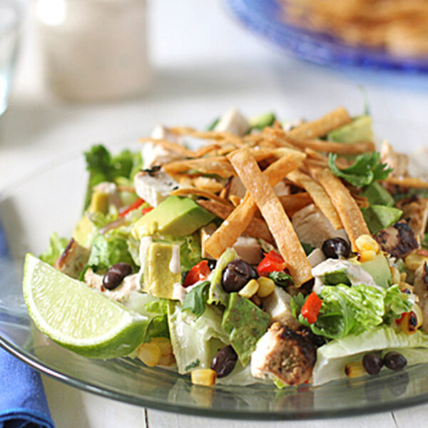 A glass plate with a salad of lettuce, black beans, corn, avocado, diced chicken, chopped red peppers, cilantro, and crispy tortilla strips, garnished with a lime wedge. A blue napkin and fork are beside the plate.