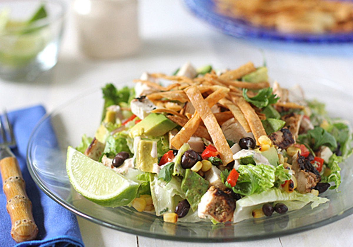 A glass plate with a salad of lettuce, black beans, corn, avocado, diced chicken, chopped red peppers, cilantro, and crispy tortilla strips, garnished with a lime wedge. A blue napkin and fork are beside the plate.