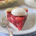 A slice of strawberry pie topped with a dollop of whipped cream sits on a white plate with a fork, with the rest of the pie and a bowl of whipped cream in the background.